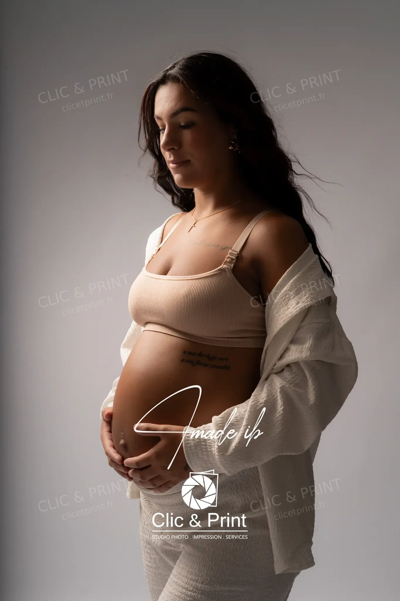 Portrait de grossesse en tenue ivoire sur fond gris, mains sur le ventre et signature Clic & Print
